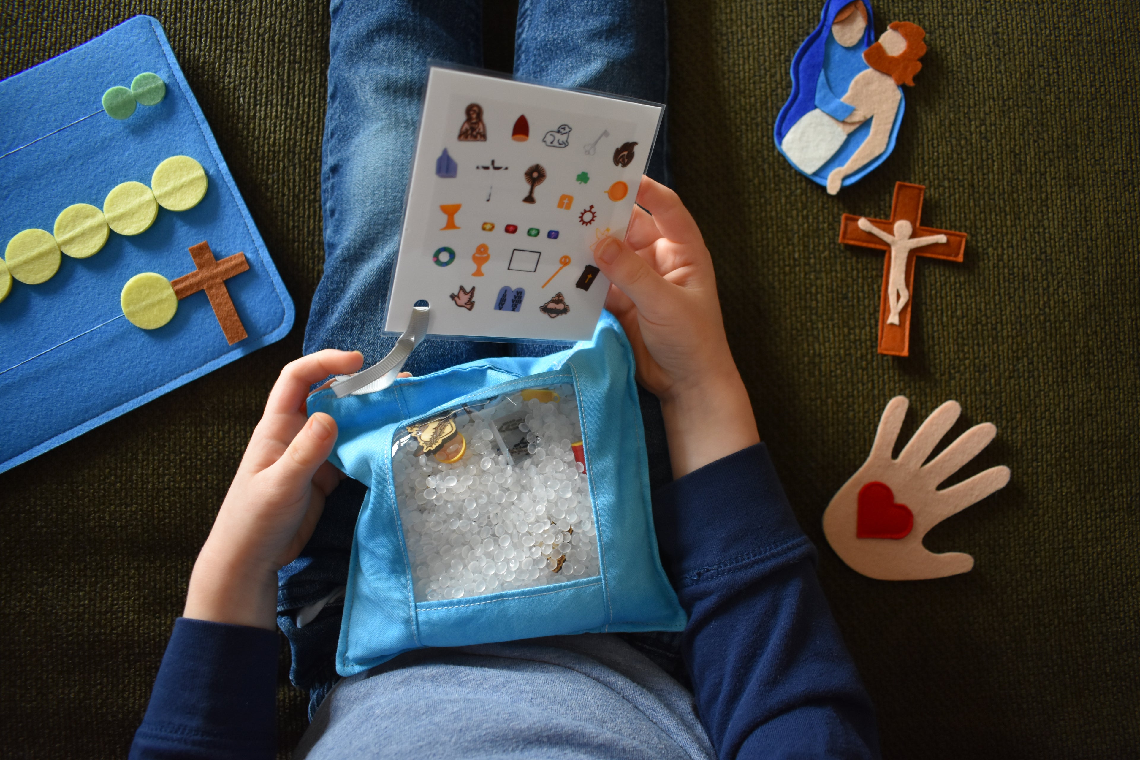Child sitting on a couch playing with Campio Kids products - Symbols of Faith I Spy bag in hand, Little Lap Rosary and Family Stations of the Cross pieces on either side of child
